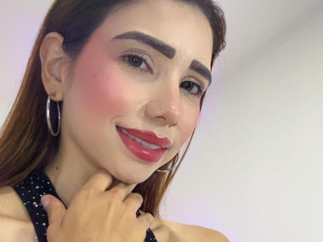 Bellasweeth ha un leggero rossore sulle guance, rossetto rosso, orecchini a cerchio e capelli castani. Tiene il collo con una mano e sorride. Il suo seno non è visibile..