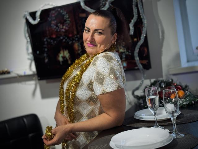 DianaMGG with pink lipstick, brown hair in a ponytail, wearing a glittery white dress and gold tinsel, smiling at the camera, holding tinsel, table with glasses in front.