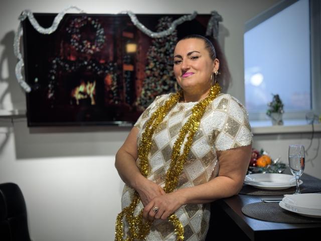 DianaMGG with pink lipstick, wearing a diamond-patterned dress and gold tinsel, standing by a table with a glass of champagne, looking confident with a slight smile.