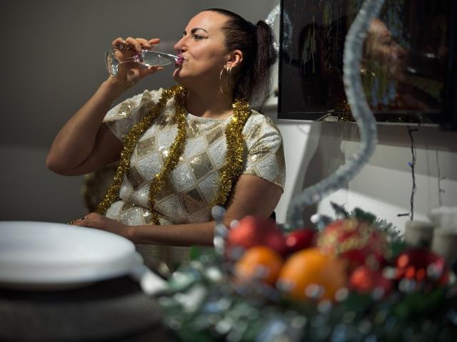 DianaMGG with dark hair in a ponytail, drinking from a glass, wearing a gold and white patterned top, and gold tinsel, standing near a festive table with ornaments.