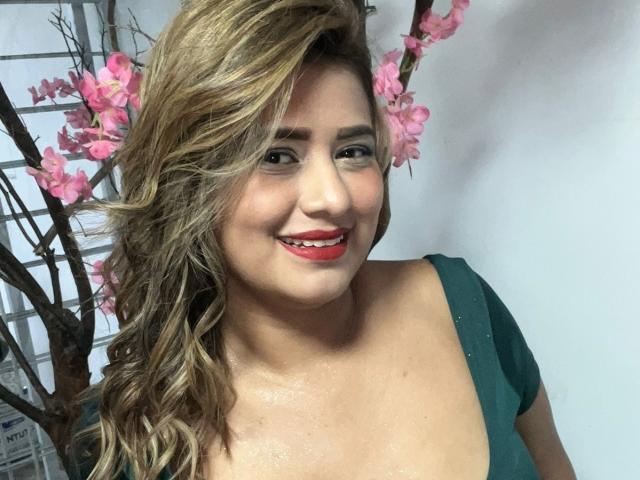 TheCookingSecret has wavy blonde hair, red lipstick, and a green dress with a low neckline, smiling at the camera with her tits slightly visible. Pink cherry blossoms in background.
