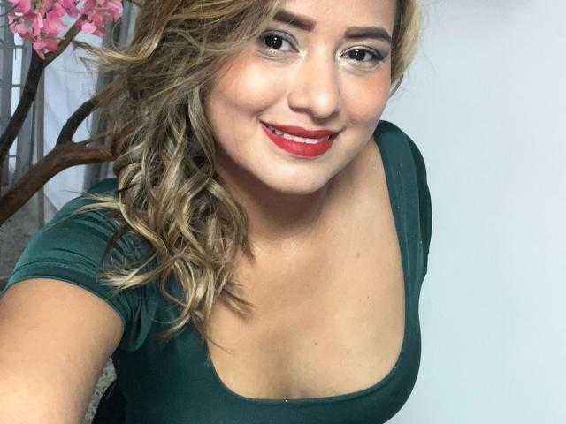 TheCookingSecret has blonde curly hair, red lipstick, and a green top showing her tits. She's smiling, with light makeup and gray eyes, taking a selfie.