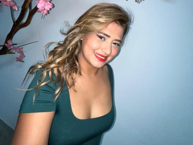 TheCookingSecret has blonde wavy hair, red lipstick, and a green top showing her tits. She's smiling with a light blue background and pink cherry blossom branches.