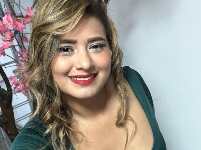 TheCookingSecret has blonde wavy hair, red lipstick, and green top, smiling with red lipstick, showing her tits slightly in a green top, against a white background.