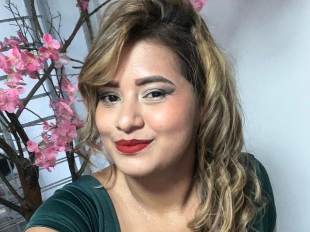 TheCookingSecret has red lipstick, wavy blonde hair, green eyes, and is wearing a green top. Pink flowers are in the background. She's smiling slightly with a subtle glow.