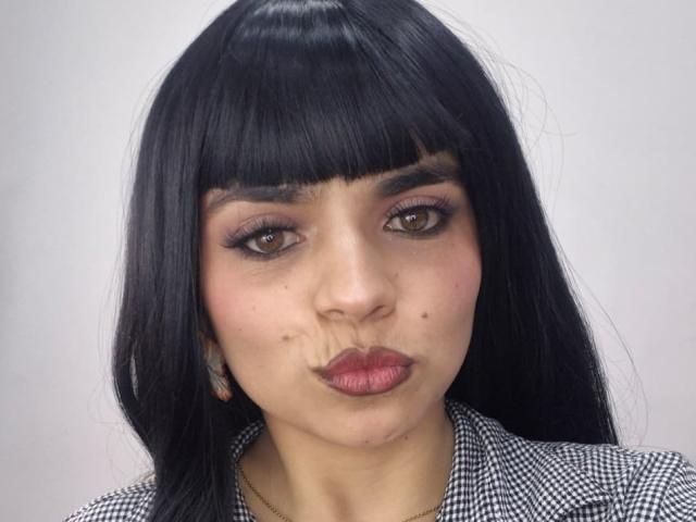 CatalinaRuiiz with black straight hair, blunt bangs, brown eyes, pink lipstick, pouty lips, small moles on her nose, wearing a black-and-white checkered shirt.
