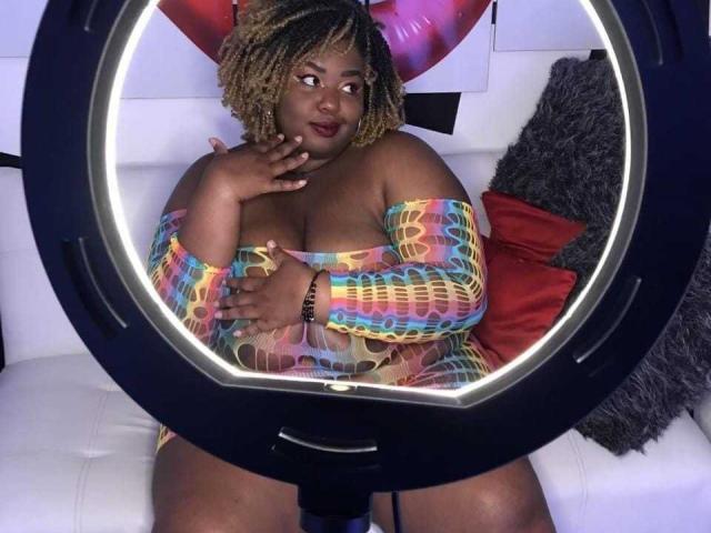 ValeryHanze with dark skin, big ass, and big boobs, wearing a colorful, off-shoulder, see-through dress, sitting in a ring light, looking to the side with a hand on her chest.