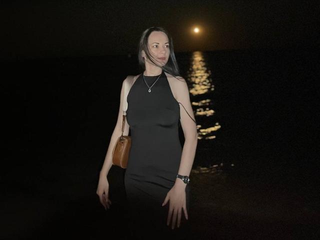 Dalaila with long dark hair, black sleeveless dress, brown handbag, silver necklace, and black wristwatch, standing by dark water with a moonlit reflection.