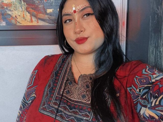 KalpanaAisha has long black hair, red lipstick, and a red and black patterned dress. She's wearing a gold chain and a jeweled forehead piece. Her tits are small.