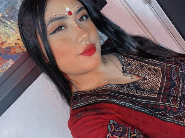 KalpanaAisha with long black hair, red lipstick, and intricate face makeup, wearing a red and black patterned top. Her ass is not visible. She's looking at the camera.