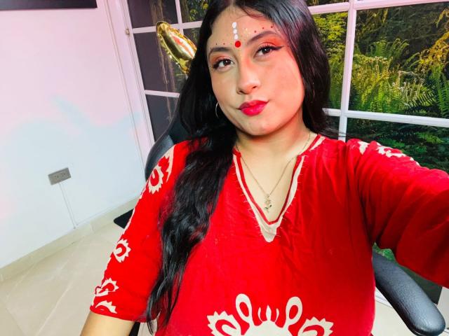 KalpanaAisha with long black hair, red dress, white floral patterns, red dot on forehead, red lipstick, gold headpiece, and necklace, sitting in chair, taking selfie.
