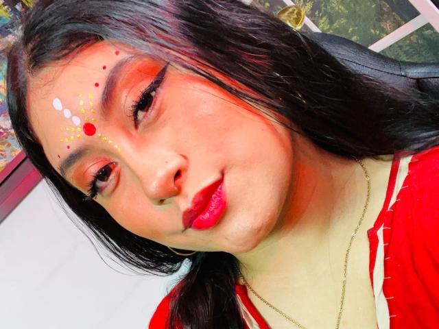 KalpanaAisha has red lipstick, dark eyeliner, and face paint with red, white, and yellow dots on her forehead. She's wearing a red top, with long black hair.