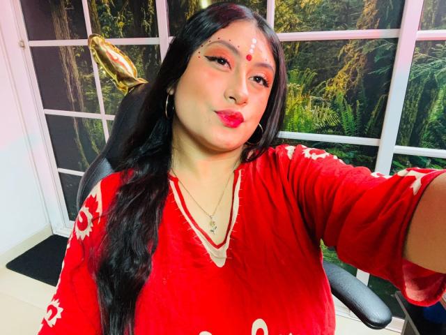KalpanaAisha, with long black hair, red lipstick, and white facial markings, pouts her glossy pink lips. She's wearing a red velvet top with white patterns, in a chair.