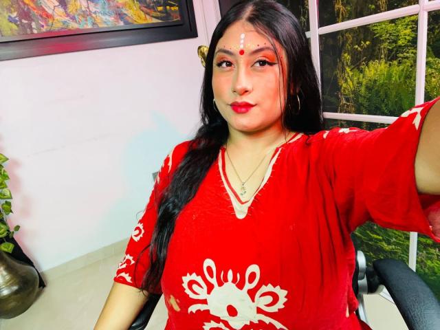 KalpanaAisha with long black hair, red lipstick, red dress with white pattern, red bindi, nose ring, and hoop earrings, holding out arm, sitting in chair.