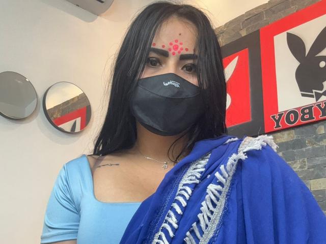 Dalia Dubey, con i capelli scuri, indossa una maglietta blu, una sciarpa blu, una mascherina nera e un punto rosso come trucco sulla fronte, mostrando un piccolo tatuaggio sul lato destro del petto..
