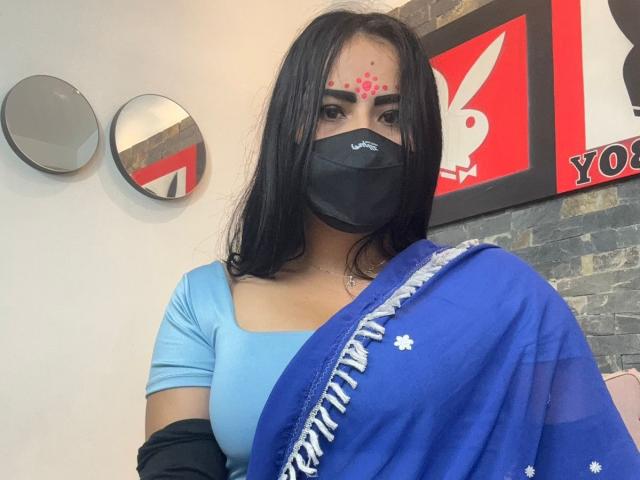 DaliaDubey with long black hair, blue sari, black mask, red forehead dots, light blue top, black sleeves, and a cute face, standing in a room. Boobs visible.