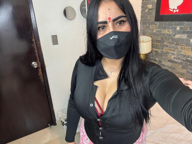 DaliaDubey with long black hair, wearing a black mask, black shirt with cleavage, red bra, and pink skirt, has a red dot on her forehead, looking at camera.