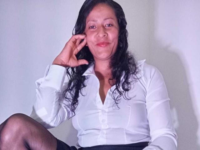 AntoHotie with curly black hair, wearing a white button-up shirt and black skirt, sitting with a relaxed smile, touching her face, showing her small tits and black pantyhose.