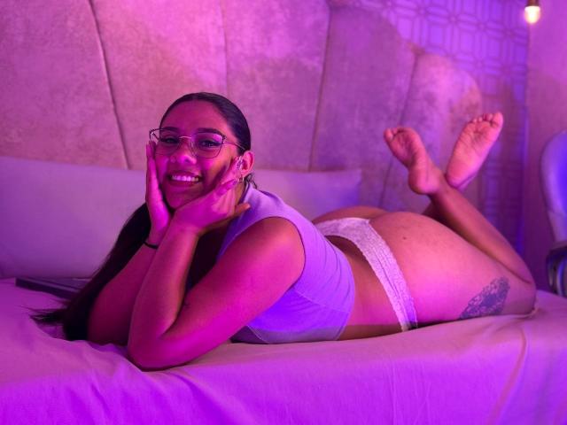 SashaSpicy lying on a bed, smiling, wearing glasses, white lace panties, and a purple top, showing her ass and a tattoo on her right thigh, under purple lighting.
