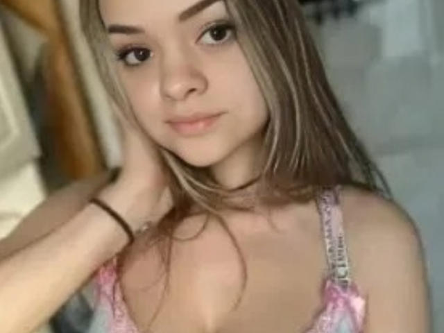 UrsulaX with long blonde hair, brown eyebrows, pink bra, slight cleavage, hand on head, subtle smile, light skin, bracelet on wrist, soft focus, slight blush on cheeks.