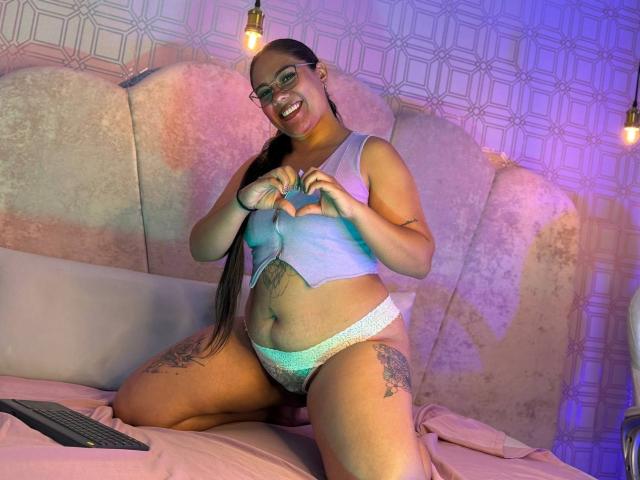 SashaSpicy, with glasses, long black ponytail, smiling, pulling up a light blue top, exposing her belly and wearing white lace panties, sitting on a bed with a keyboard.