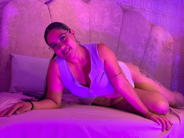 SashaSpicy, lying on bed, wearing white crop top, revealing boobs, and white underwear, with purple lighting, glasses, tattoos, and a cute smile. Her ass is partially visible.