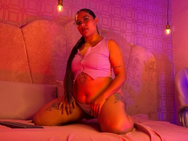 SashaSpicy with long brown ponytail, pink crop top, and white panties, kneeling on bed, showing tattoos on thighs, with hand on pussy, pink and purple lighting.