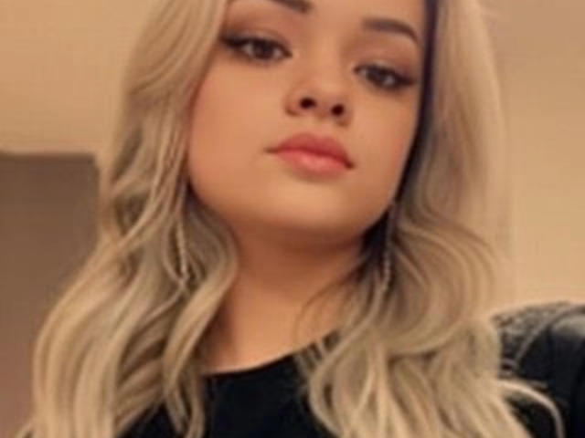UrsulaX with blonde wavy hair, full pink lips, and subtle makeup, wearing a black top, has a soft, pretty face, and is looking slightly to the side.