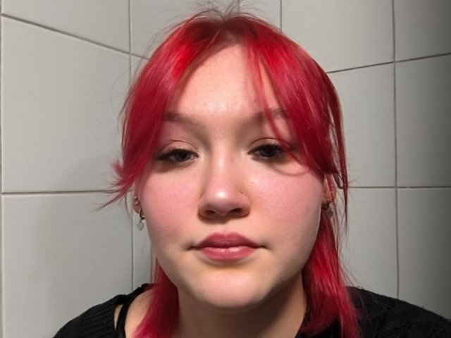 CloraFlippin has bright red hair, fair skin, small nose piercing, subtle makeup, and black shirt, looking straight into the camera with a slight smile.