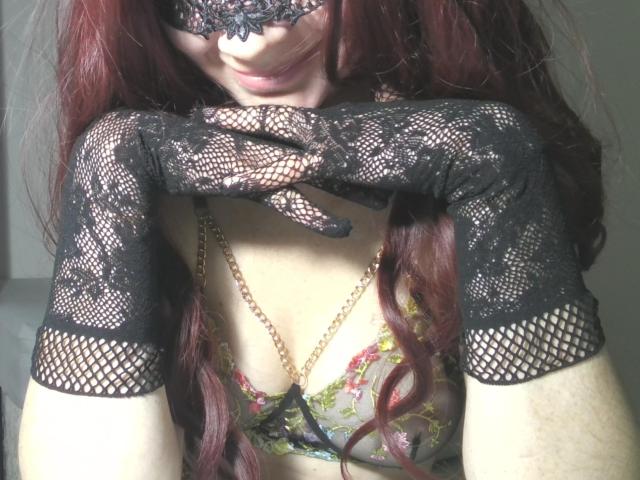 Close-up of AromaLove with long, wavy red hair, wearing a black lace mask, black lace gloves, and a floral lace bra with a gold chain. AromaLove's hand is raised to her chin, fingers touching her mask. The lace gloves have a fishnet pattern at the cuffs. AromaLove's pale skin contrasts with the dark lace. The image focuses on the upper body, emphasizing the intricate lace patterns and AromaLove's facial expression. The lighting is soft, highlighting the textures of the lace and hair.