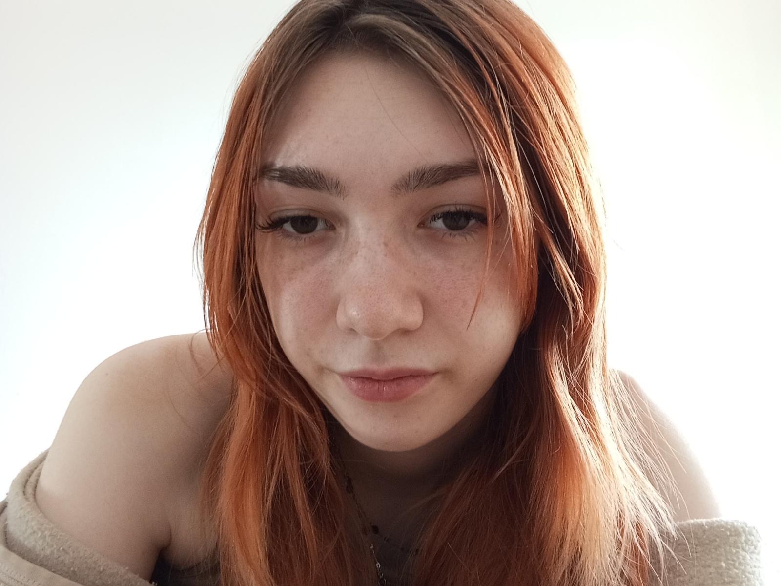 PaulDursch has orange hair, freckles, and is looking down with a neutral expression, wearing a beige off-shoulder top, exposing her shoulders. Her small tits are partially visible.