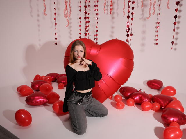 Margarita Kis con un globo rojo en forma de corazón, una blusa negra y pantalones vaqueros grises, arrodillada sobre un suelo blanco, rodeada de globos rojos, con un dedo en los labios, luciendo seductora, piel clara, cabello rojo..