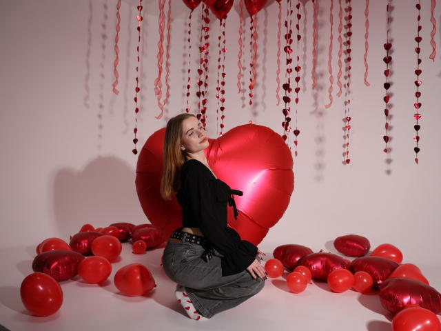 MargaritaKis with red heart balloons, kneeling, wearing black top, gray pants, and red socks, surrounded by red balloons, red heart-shaped balloon, and hanging red hearts.
