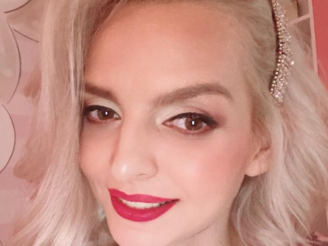 DouceAmelia's face with blonde hair, red lipstick, and brown eyes, wearing a diamond hairpiece, smiling subtly. Her makeup is subtle, highlighting her natural beauty.
