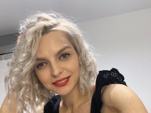 DouceAmelia has curly white hair, red lipstick, and black lace dress, smiling at the camera with brown eyes. She's holding her hair up, showing off her face.