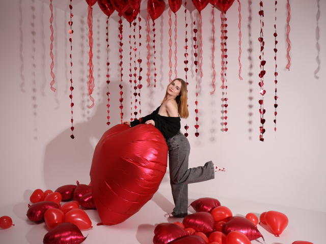 MargaritaKis with red heart balloon, black off-shoulder top, gray pants, standing on one leg, surrounded by red heart balloons and hanging red hearts, smiling.
