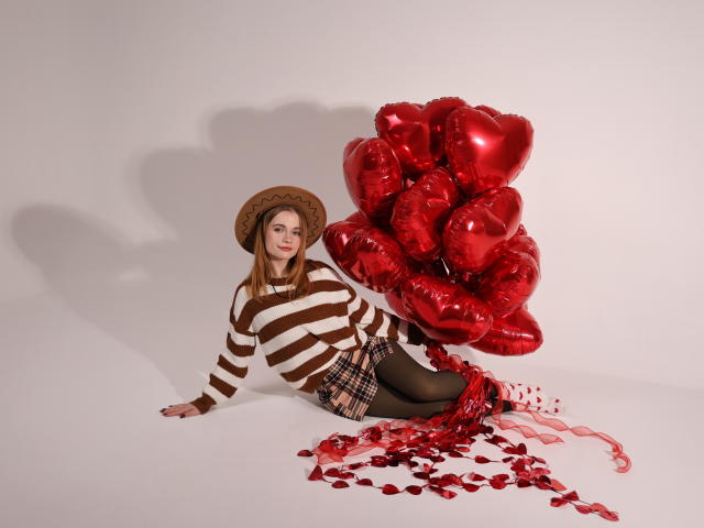 Margarita Kis con un suéter a rayas marrón y blanco, falda de cuadros, medias negras, sombrero, globos rojos con forma de corazón y pétalos de rosa roja esparcidos, sentada en el suelo blanco, sonriendo..
