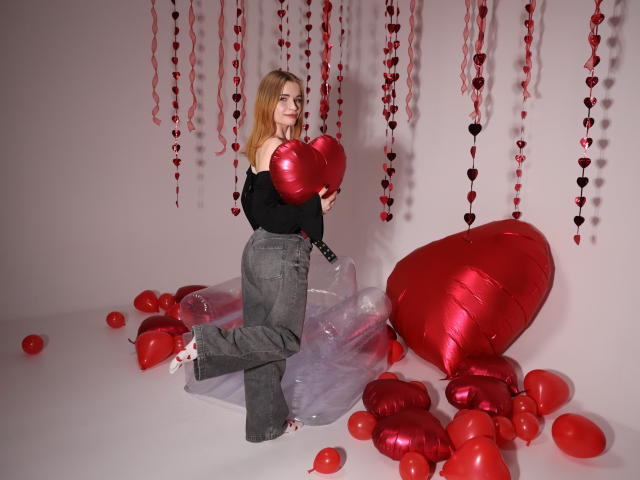 MargaritaKis with long blonde hair, black off-shoulder top, gray jeans, holding a red heart, kneeling on transparent plastic, surrounded by red balloons and heart-shaped decor.