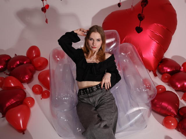 MargaritaKis with small tits, wearing a black off-shoulder top and gray pants, sits in a transparent inflatable chair, surrounded by red heart balloons and shiny heart decorations.