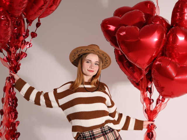 MargaritaKis con cappello marrone, maglione a righe e gonna a quadri, che tiene palloncini a forma di cuore rossi, sorridente, in piedi, nastri rossi, capelli biondi, carina, sfondo bianco..