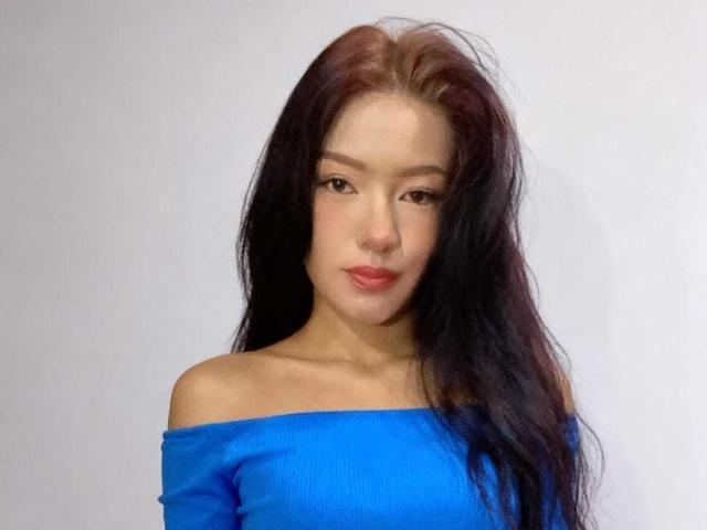 IvorySelene with long black hair, blue off-shoulder top, and red lips, looking down with a slightly serious expression, showing her bare shoulders and upper chest.