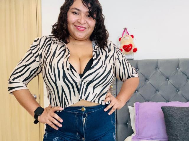 KarlaWild with curly black hair, smiling, wearing a zebra-striped top tied below her big boobs, black bra visible, blue jeans, black wristwatch, standing in a bedroom.