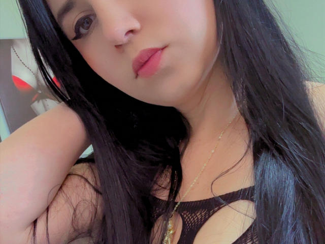 Kkatalina with long black hair, wearing a black mesh top showing her tits, has pink lips, a gold necklace, and a nose ring, looking at the camera.