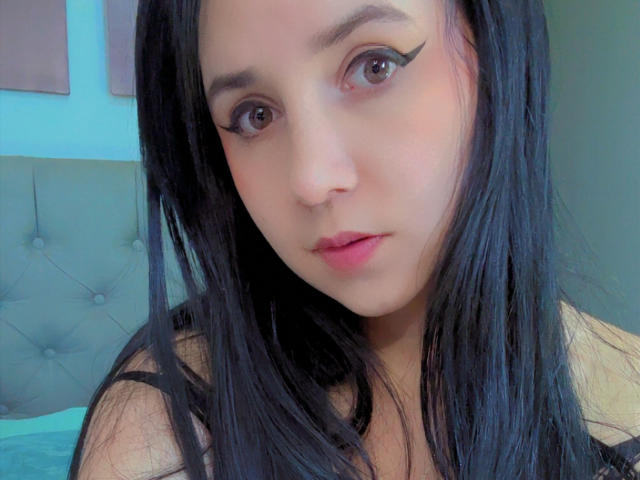 Kkatalina has black hair, winged eyeliner, pink lips, and a cute face. She's looking at the camera with a neutral expression, wearing a black top.