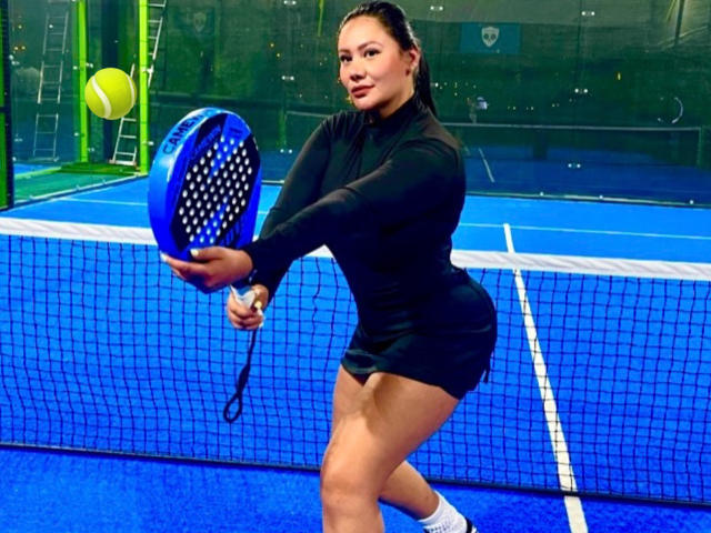 AlesandraMalory, a woman with tan skin and dark hair, is mid-swing with a blue and black tennis racket. She wears a black long-sleeve top and black shorts. She's positioned in the center, with a bright blue tennis court and a net behind her. Her racket is angled upward, and a yellow tennis ball is in the air to her left. She's also wearing white socks. The image is brightly lit with a focus on her athletic form.