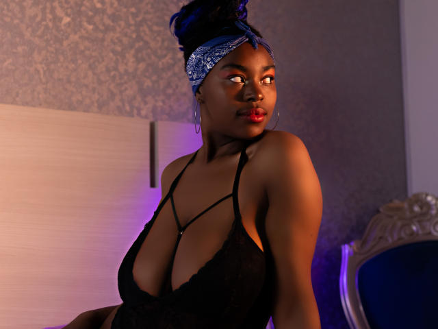 KrisMond, dark-skinned, wearing black lace lingerie, deep cleavage, blue headscarf, looking to the side, red lipstick, sitting in a room with purple and orange lighting.