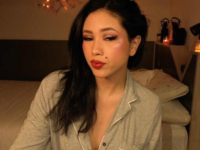 NashiroSakuraii with long black hair, wearing a gray button-up shirt, pouting with red lipstick, a small mole on her cheek, and pink eye makeup in a warmly lit room.