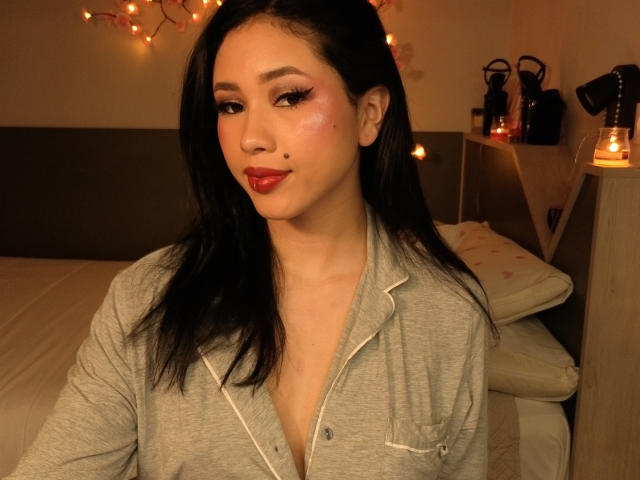NashiroSakuraii with black hair, red lipstick, and a grey button-up shirt, showing a beauty mark on her right cheek, sitting in a warmly lit bedroom.