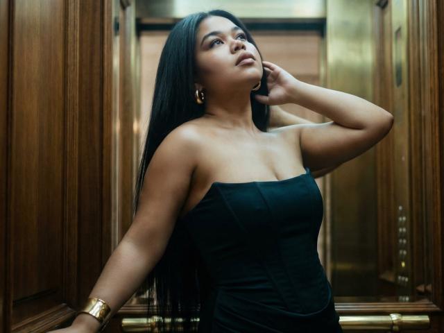 SunshineFlow with long black hair, wearing a strapless black dress, gold bracelet, and earrings, standing in a wooden elevator, looking up with one hand on her neck.