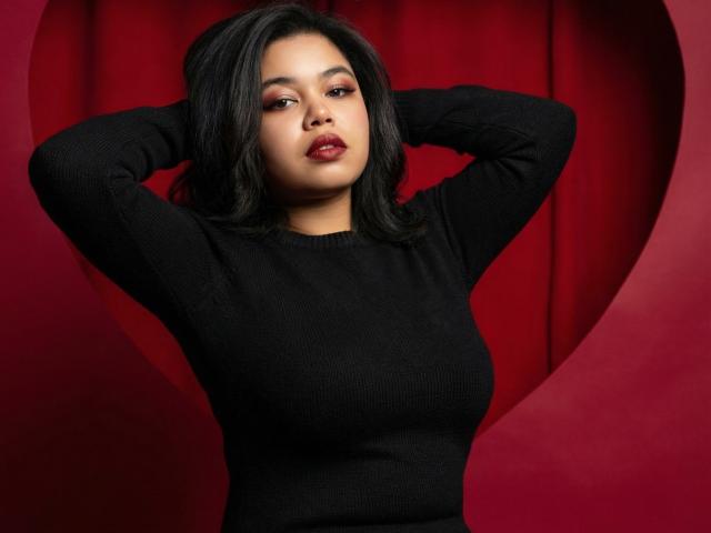SunshineFlow with dark hair, wearing a black sweater, arms raised, hands behind head, red lipstick, looking at camera, medium skin tone, medium-sized tits, confident expression, red background.